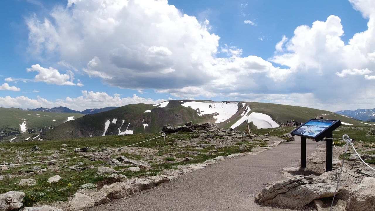 Wide ground-level view in Winter Park, Colorado with a paved overlook path, low rocks, rope barriers, distant green ridges, snow patches, large clouds, and no visible people.