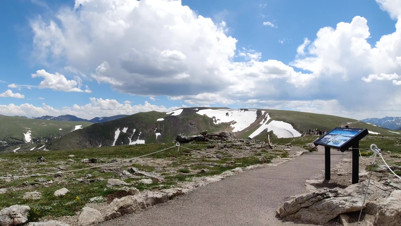A second wide view in Winter Park, Colorado frames the paved path and interpretive sign closer to center, with rocky foreground, rope barrier, rolling ridges, snow patches, and no visible people.