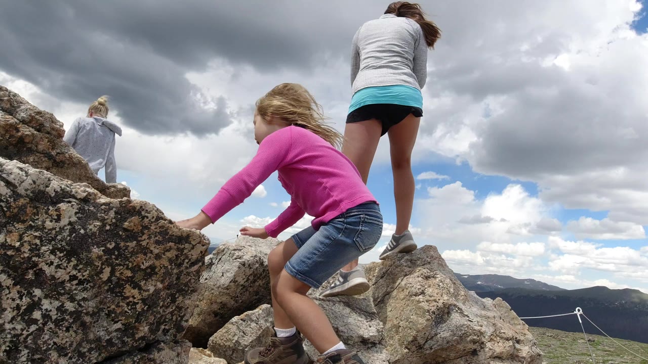 Girl in pink climbing rocks near two other people under cloudy sky.