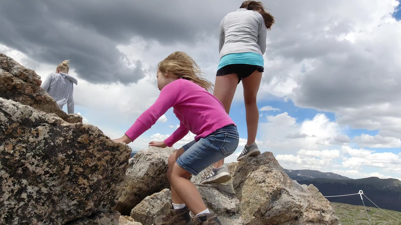 The girl continues climbing across lichen-covered rocks in Winter Park, Colorado, with an adult and another child positioned above her against the mountain horizon.