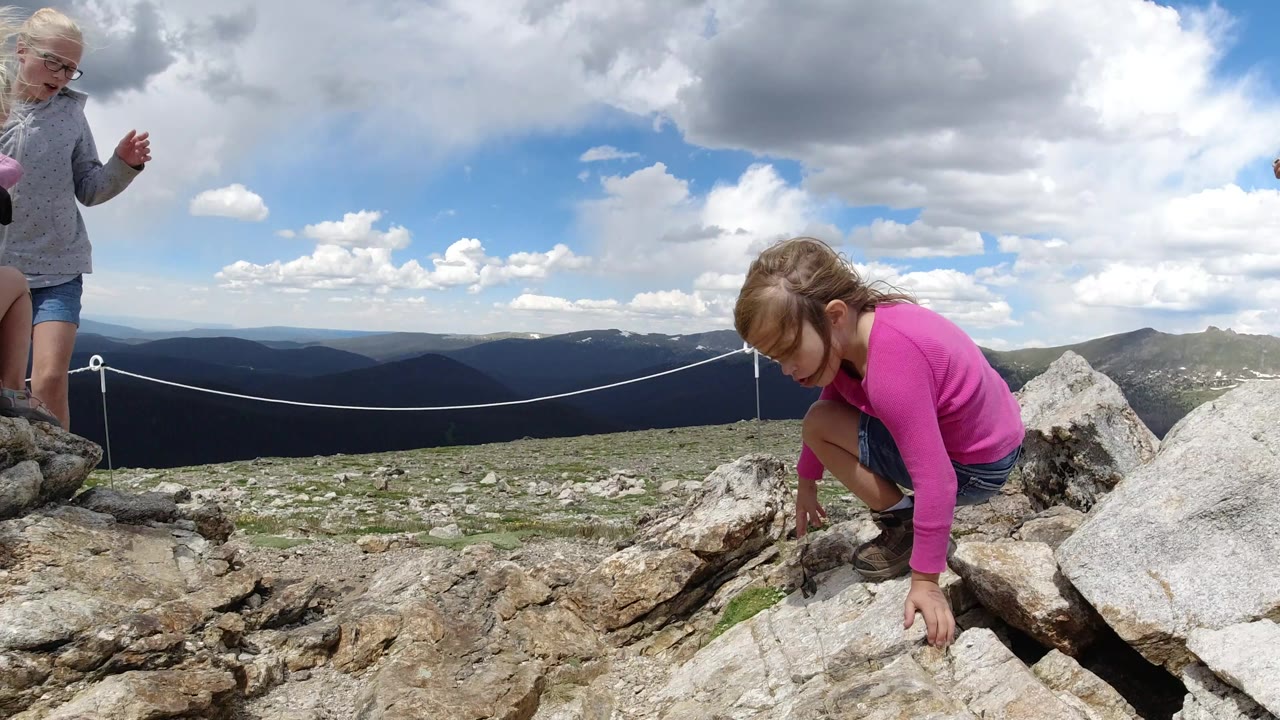 A child in a bright pink top crouches and reaches toward the rocks while a blond child in gray stands nearby at a Winter Park, Colorado overlook with distant mountains and clouds.