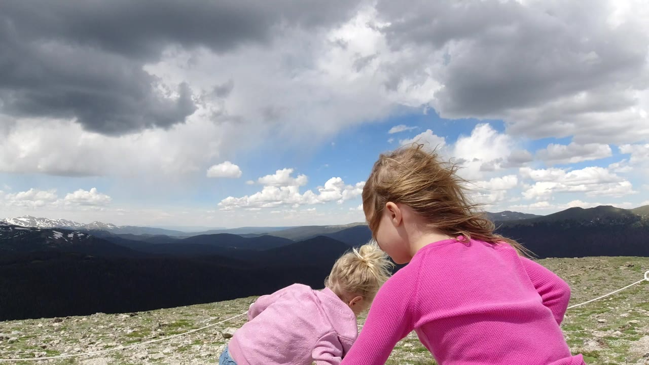 Two girls crouch on rocky mountain ground under large clouds.