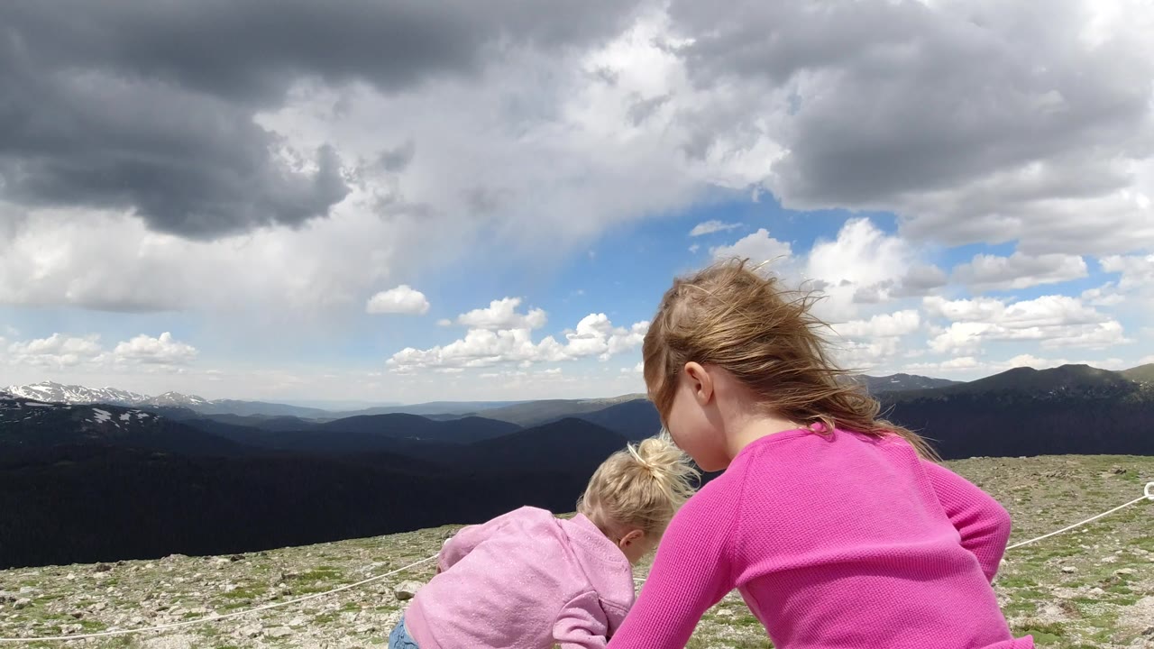 The girls lean forward on a rocky slope in Winter Park, Colorado, while a rope line and layered mountain ridges stretch across the background.
