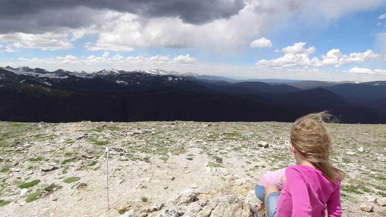 The child remains near the lower right foreground on pale rocky terrain in Winter Park, Colorado, with layered ridgelines and a dark cloud bank overhead.