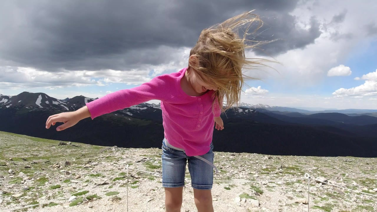 Blond girl in a bright pink shirt stands on pale rocks in Winter Park, Colorado, with windblown hair and dark mountain ridges behind her.
