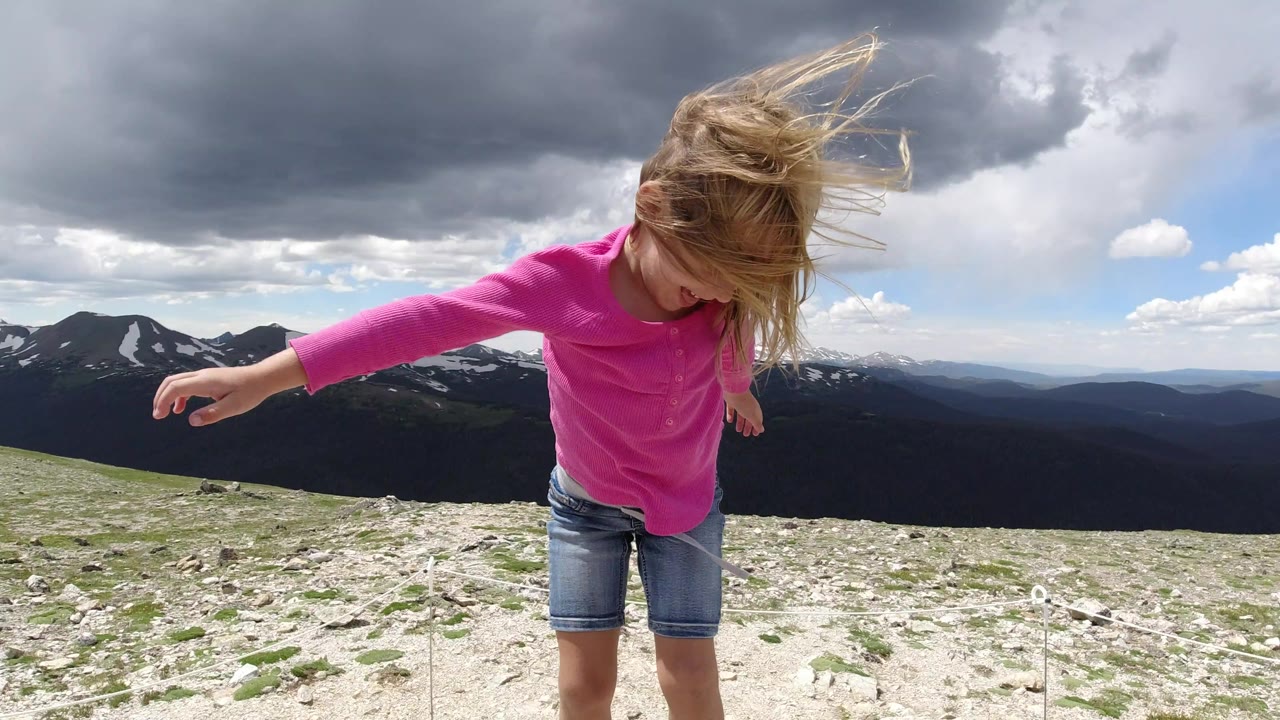 The child leans forward on a rocky slope in Winter Park, Colorado, while wind lifts her blond hair under heavy gray clouds.