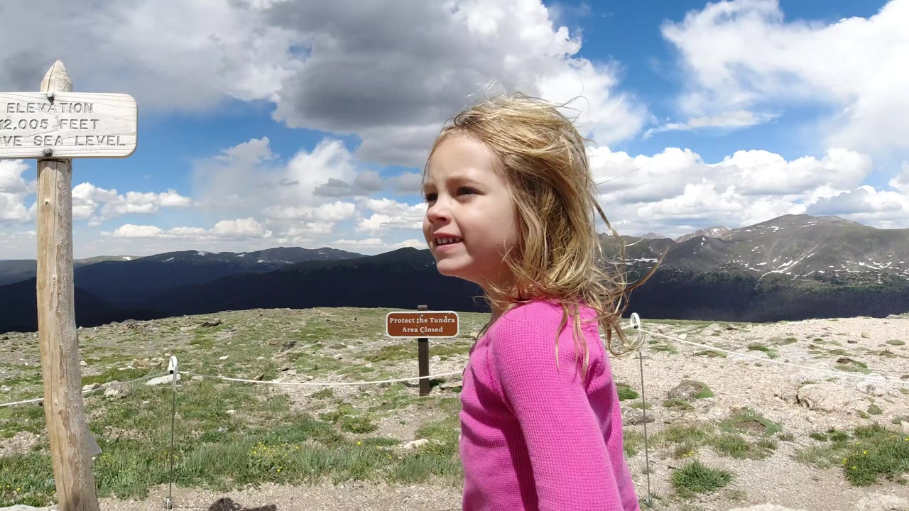 The girl remains near the foreground signs at the Winter Park, Colorado overlook, facing across rocky tundra toward layered mountain ridges under large white clouds.