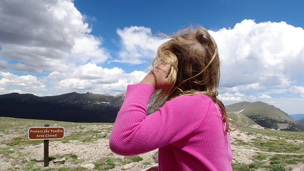 A young girl in a pink top stands in the foreground at a Winter Park, Colorado mountain overlook, with a tundra closure sign, rope line, rocky ground, clouds, and distant peaks behind her.