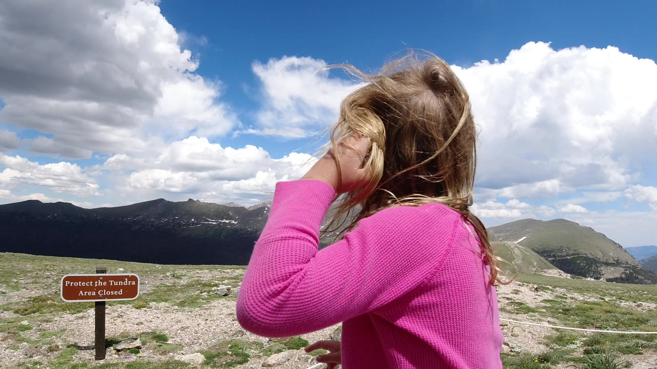 The girl turns slightly with one hand near her face at the Winter Park, Colorado overlook, while the tundra closure sign sits lower in frame and mountains stretch across the horizon.