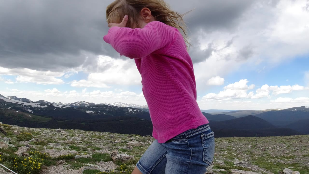 A child in a bright pink shirt crosses the foreground in Winter Park, Colorado, with rocky alpine ground, distant snow-patched mountains, and heavy gray clouds behind them.