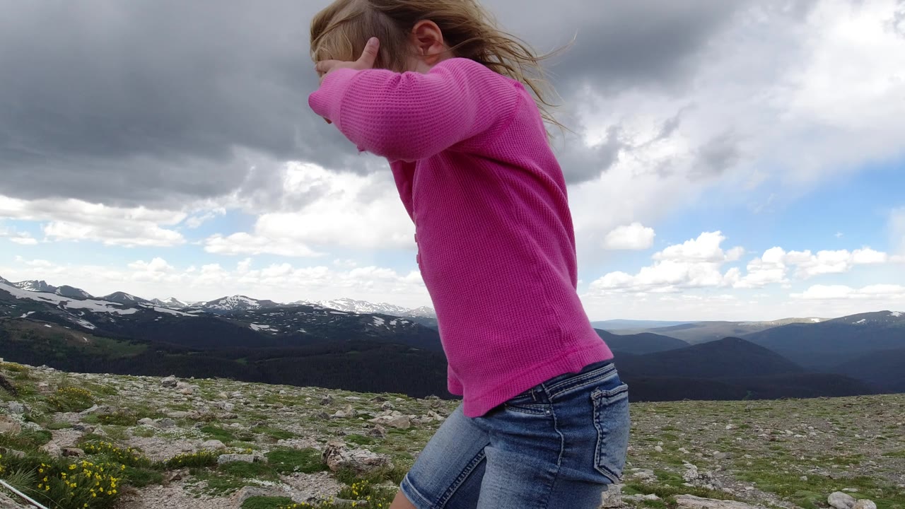The child remains near the left foreground in Winter Park, Colorado, walking across rocky terrain while layered mountain ridges and cloud breaks fill the background.