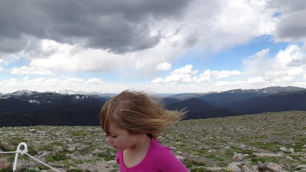 A young girl in a pink shirt appears at the lower left of a Winter Park, Colorado mountain view with rocky ground, rope posts, distant ridges, and heavy clouds overhead.