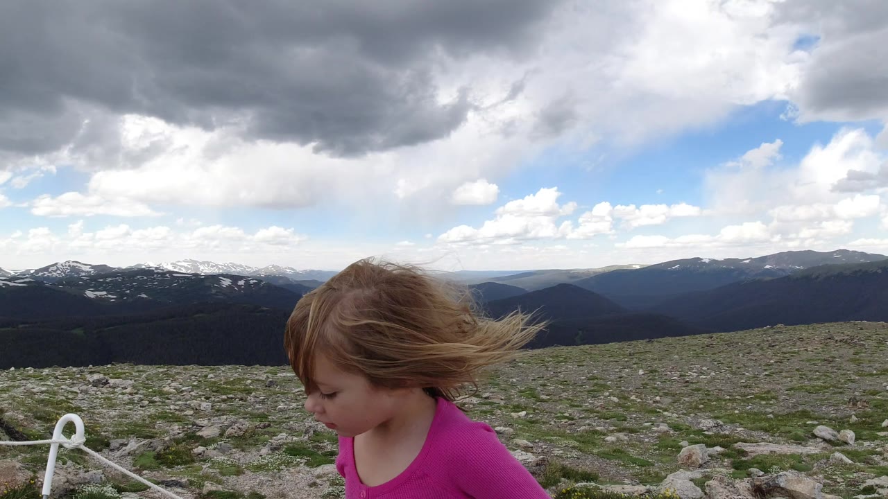 The girl moves farther into the frame in Winter Park, Colorado, looking down as wind pushes her hair across a rocky foreground and layered mountain ridges sit behind her.