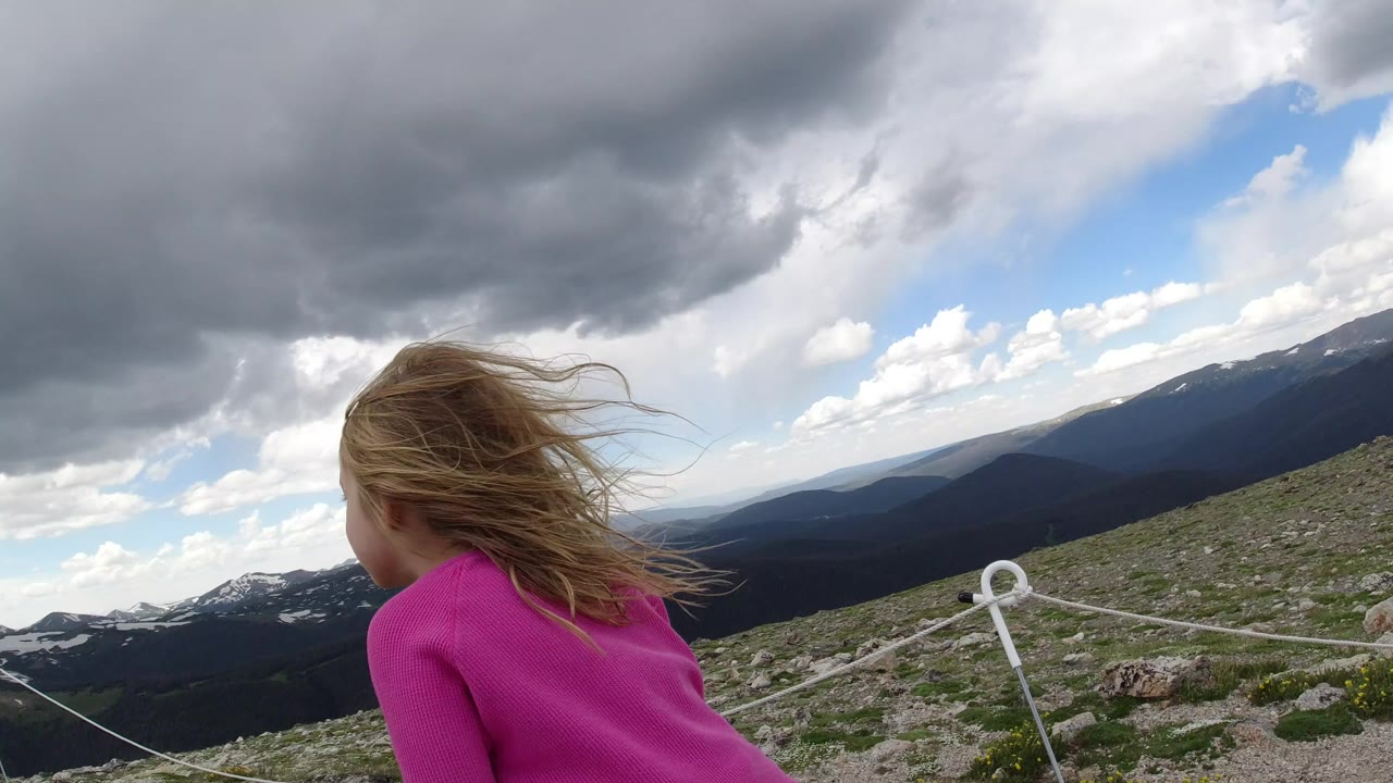 A blonde child in a bright pink shirt faces away near a white rope barrier on a rocky Winter Park, Colorado mountain overlook under dark clouds.
