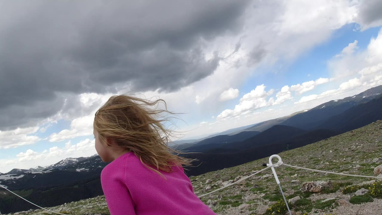 The child remains turned away as wind pushes her hair across the frame, with rope posts, rocky ground, and distant ridges visible in Winter Park, Colorado.