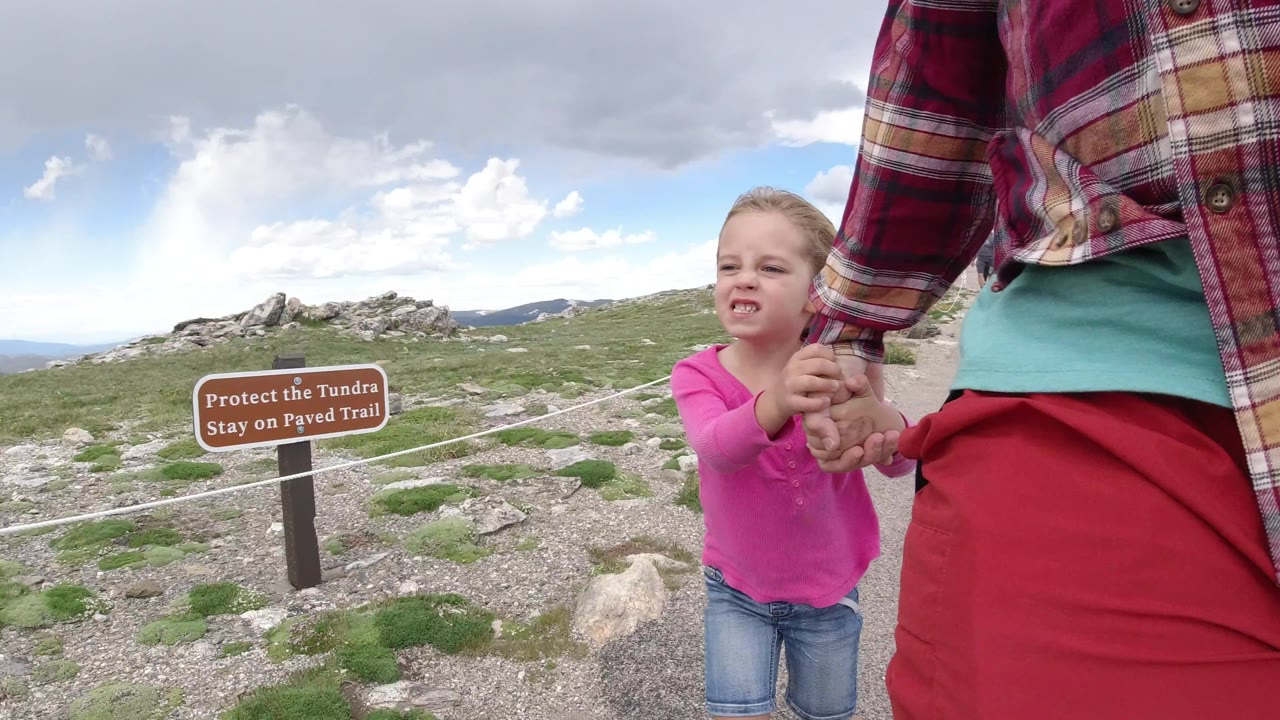 The child looks upward while holding the adult's hand on the Winter Park trail, with the tundra sign left of center and open rocky ground in the background.