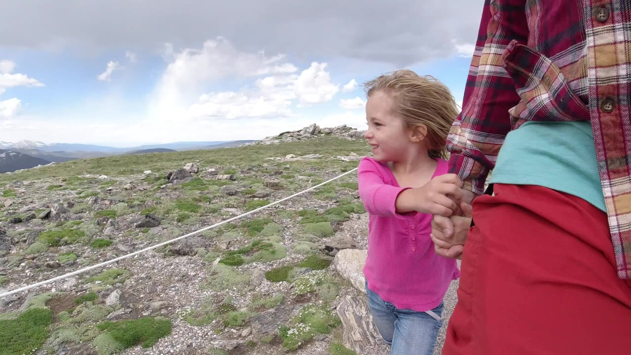 At the start of the Winter Park, Colorado clip, a young girl in a pink shirt holds an adult woman's hand beside a rocky path with low alpine plants and distant ridges.