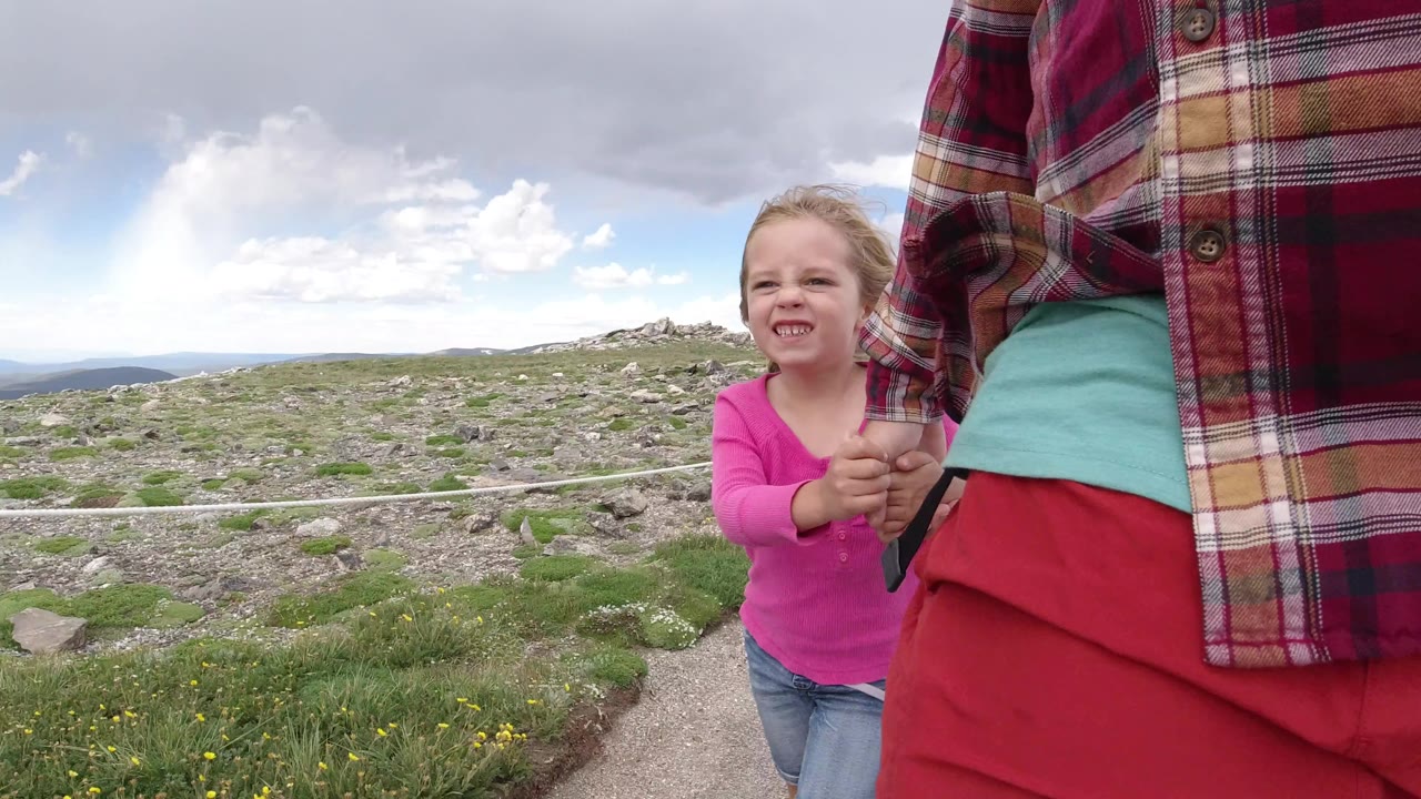 At 0:01, the girl faces forward while holding the cropped adult's hand, with the rope barrier crossing the rocky Winter Park, Colorado landscape in the background.