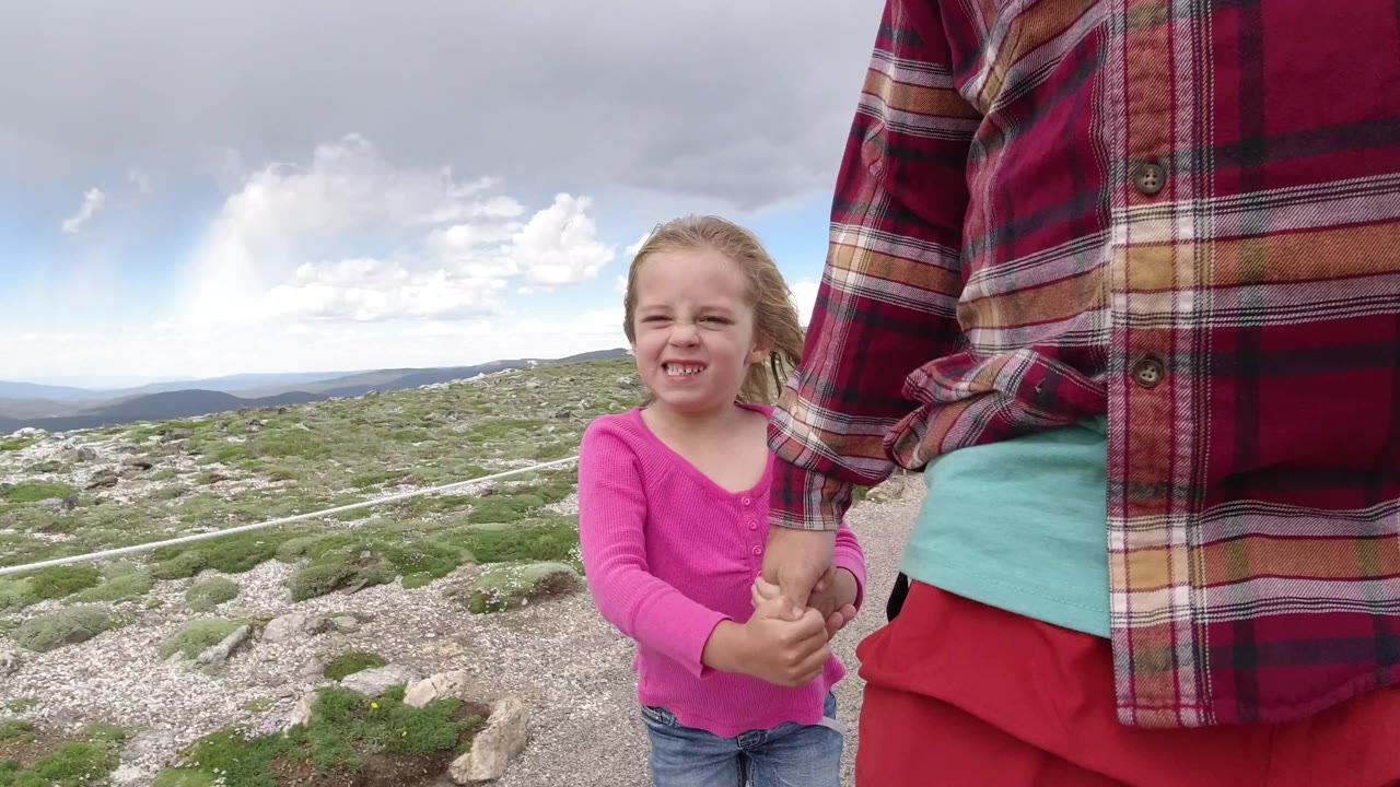 The girl faces forward while holding an adult's hand, with a pale roped path, rocky tundra, and distant ridgelines visible in Winter Park, Colorado.