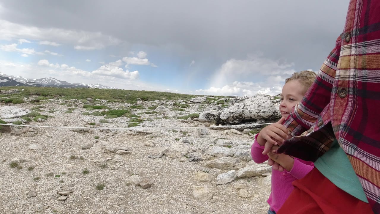 A blond child is partly obscured by an adult's plaid shirt in Winter Park, Colorado, with pale rocks, scrub vegetation, and snow-streaked mountains in the background.