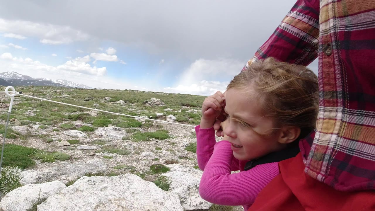 In Winter Park, Colorado, a young girl in a pink top holds an adult's hand while walking past rocks, low plants, rope trail edging, and distant snow-streaked mountains.