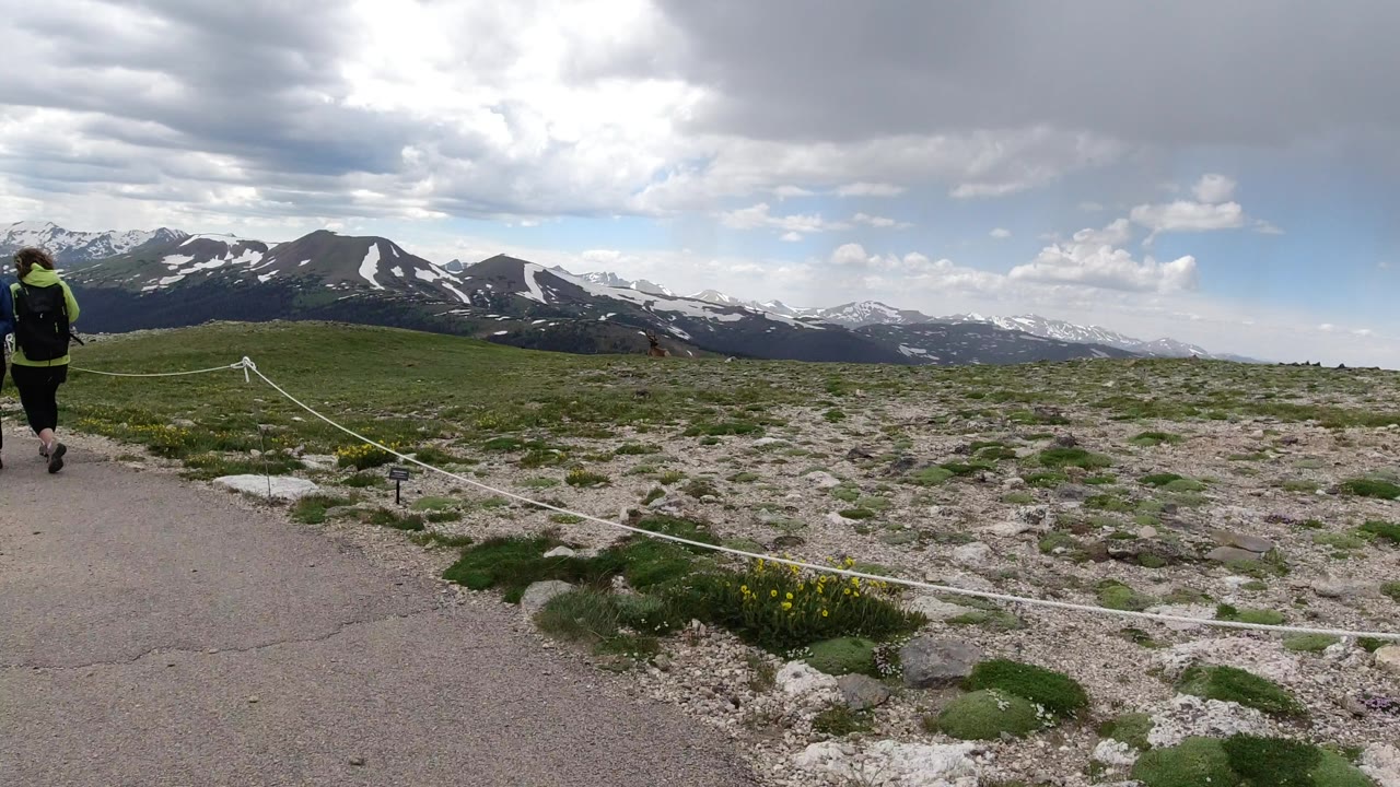 Person walking on a paved mountaintop path beside a roped boundary and distant snowy mountains.