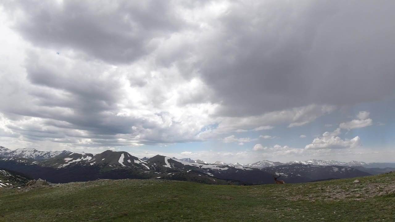 Wide Winter Park, Colorado lookout view with low green grass, distant snow-patched mountain ridges, a gray cloud ceiling, and no visible people.