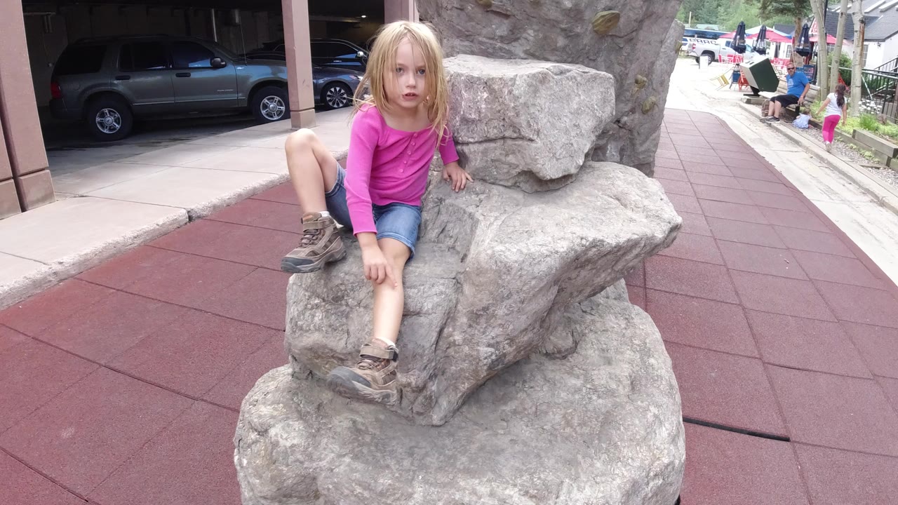 Blonde girl sitting on a gray rock in a public plaza.
