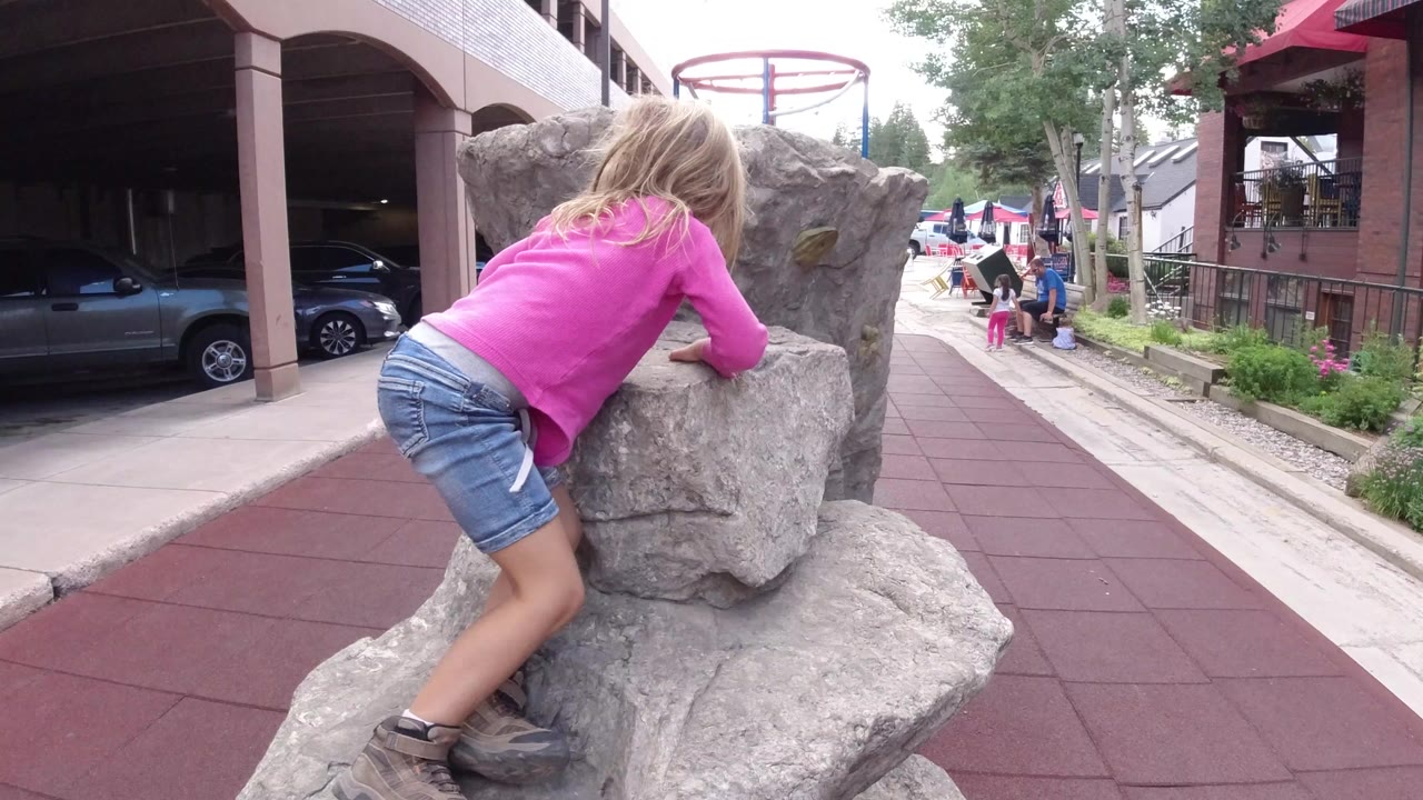 Girl in a pink shirt climbs onto a gray rock beside a paved walkway.
