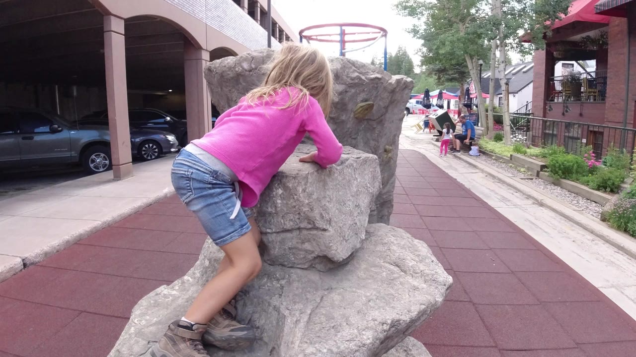 In Winter Park, Colorado, the girl braces herself against the gray rock, with one leg on the lower stone and the walkway, parked car, building columns, and distant people in the background.