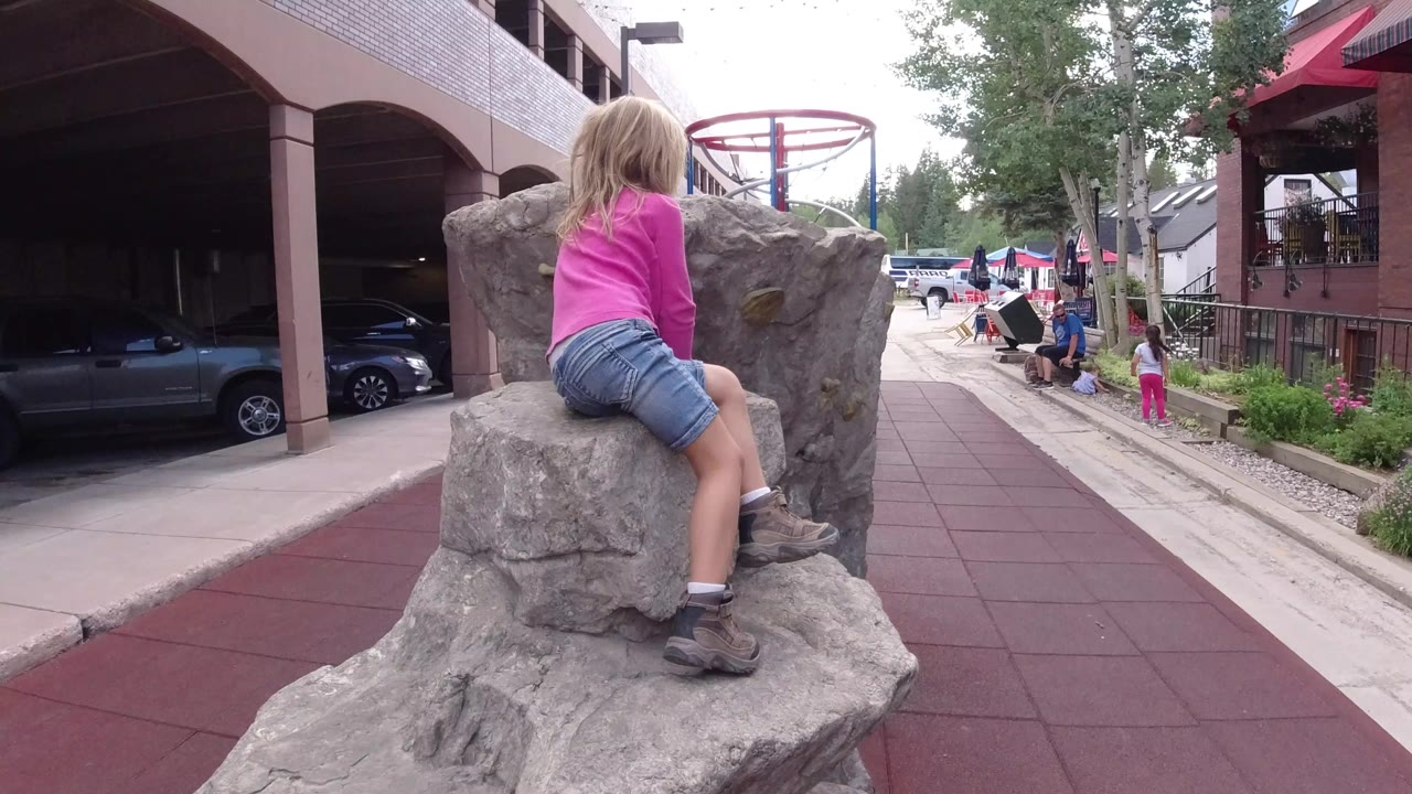 A blond young girl in a pink shirt sits on a gray artificial boulder in a Winter Park, Colorado playground, with a covered parking structure and a red play surface behind her.