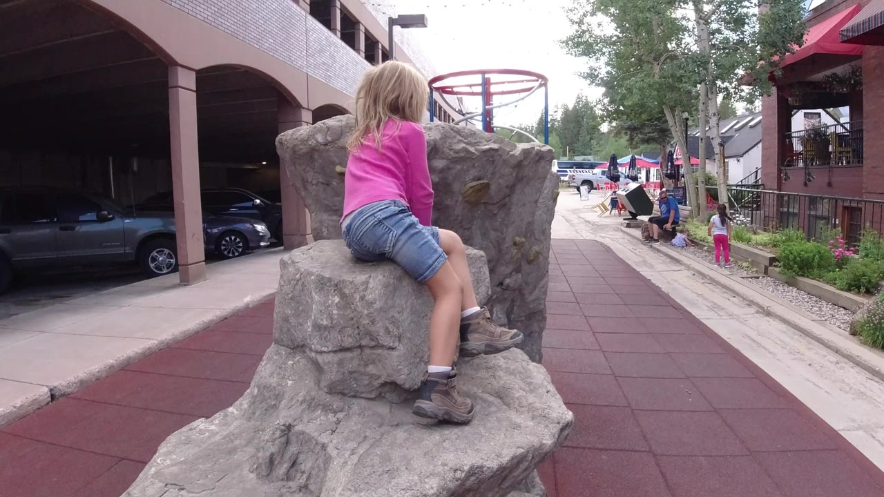The girl remains seated on the rock feature, turned slightly away from camera, while people and vehicles appear deeper in the Winter Park playground setting.