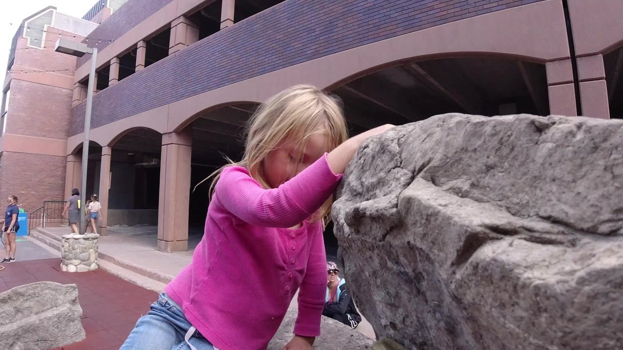 A blonde girl in a pink shirt leans forward against a large gray rock in Winter Park, Colorado, with a brick building and a few people behind her.