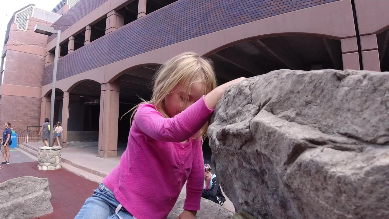 The girl is partly obscured by the rock as she grips the upper edge, with the Winter Park plaza and building columns visible in the background.