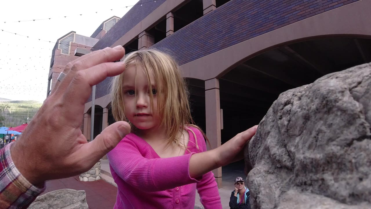 In Winter Park, Colorado, the girl looks toward the camera as an adult hand stays raised in the foreground and the boulder fills the right side of the frame.