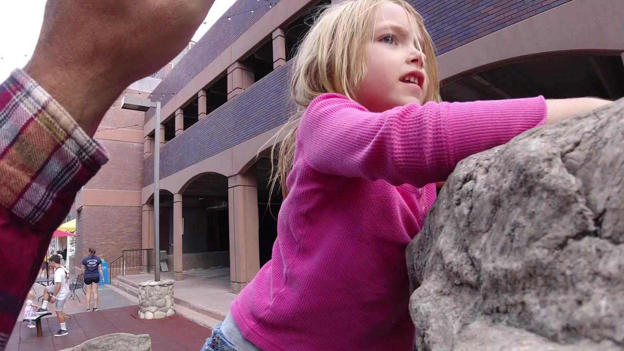 Blonde child in a pink sweater climbs a gray stone monument beside an adult arm.