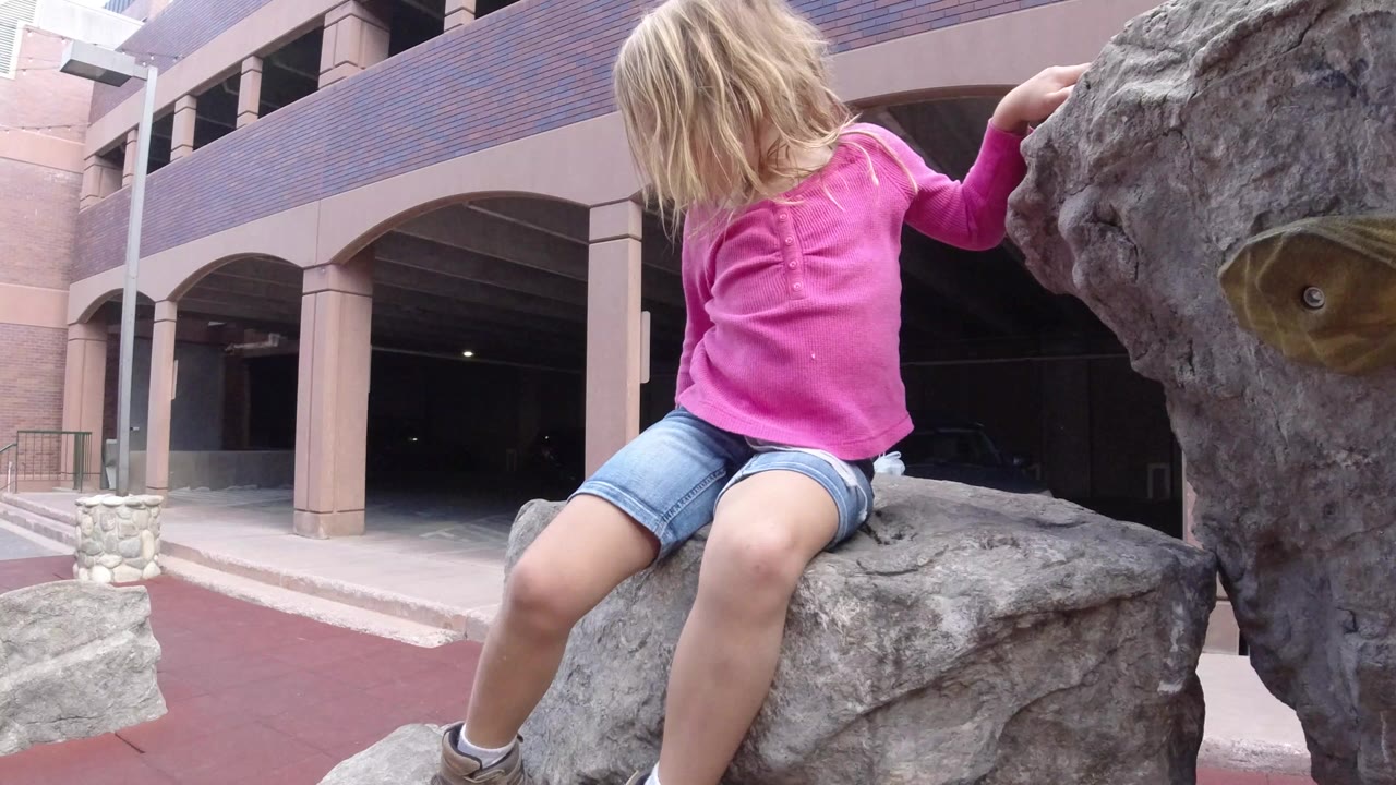 Young blond girl sitting on a gray rock-climbing statue beside a building entrance.