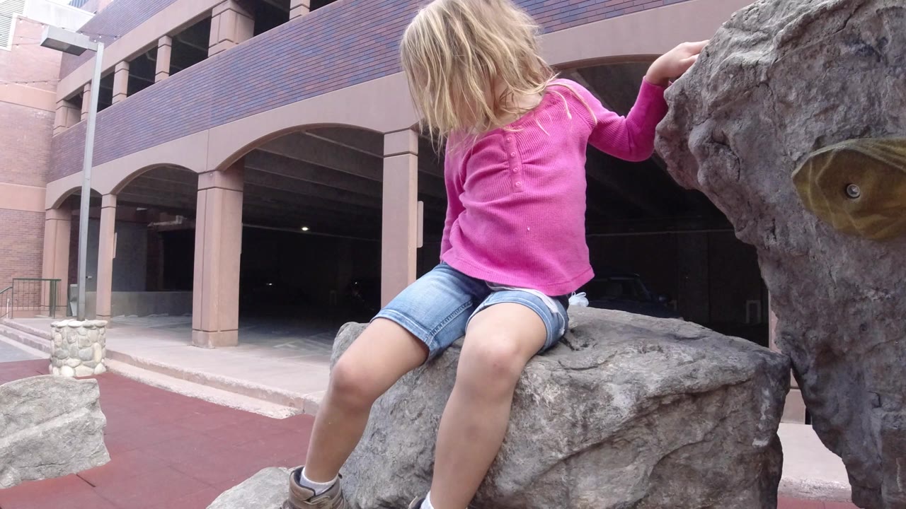 The child remains seated on the gray climbing statue in Winter Park, Colorado, while a person walks near the arched building entrance in the background.