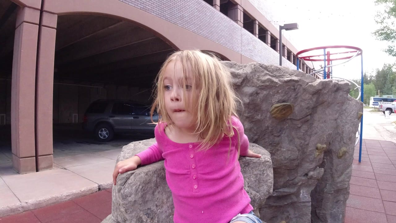 A young blonde girl in a pink shirt grips a gray stone feature in Winter Park, Colorado, with building arches and a parked vehicle behind her.