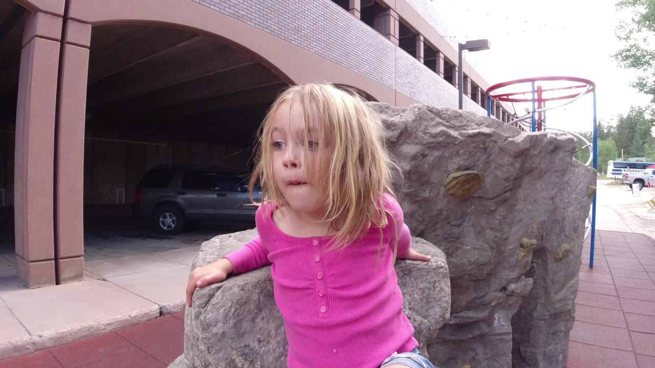 The girl remains beside the large gray boulder in Winter Park, Colorado, looking to the side while a covered parking area and sidewalk sit behind her.