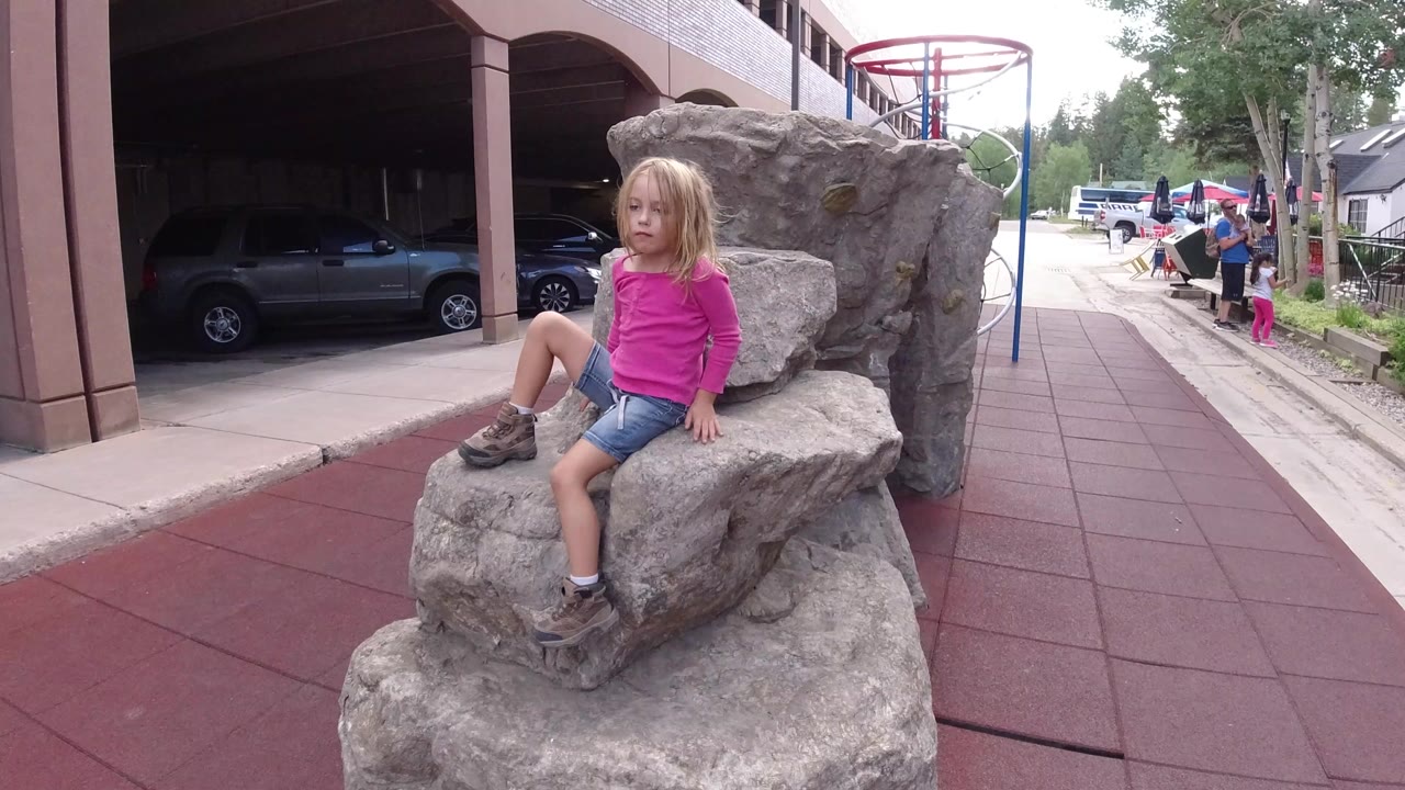 A child in a pink top sits on a rough stone chair in Winter Park, Colorado, with parked cars, columns, trees, and pedestrians in the background.