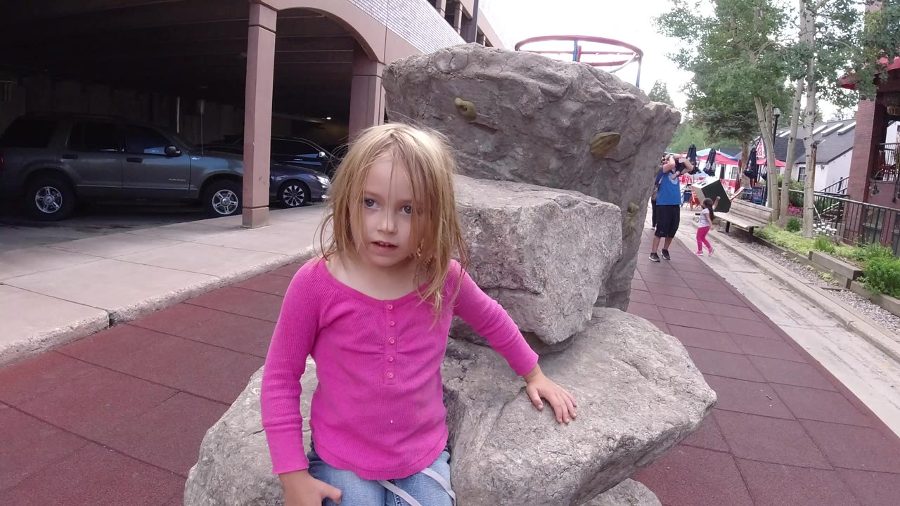 The child faces the camera from beside the artificial rock formation in Winter Park, Colorado, while a parked SUV and people on the walkway remain visible in the background.