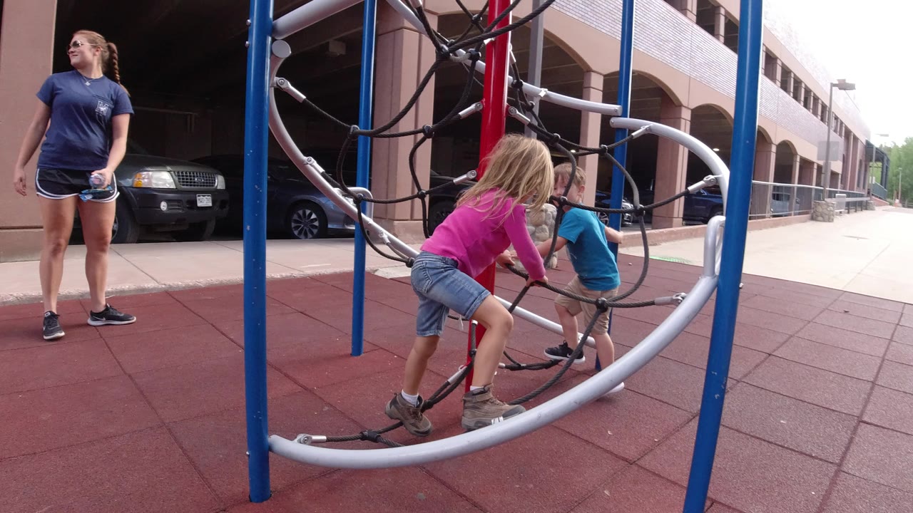 Children climbing rope playground equipment with an adult nearby.