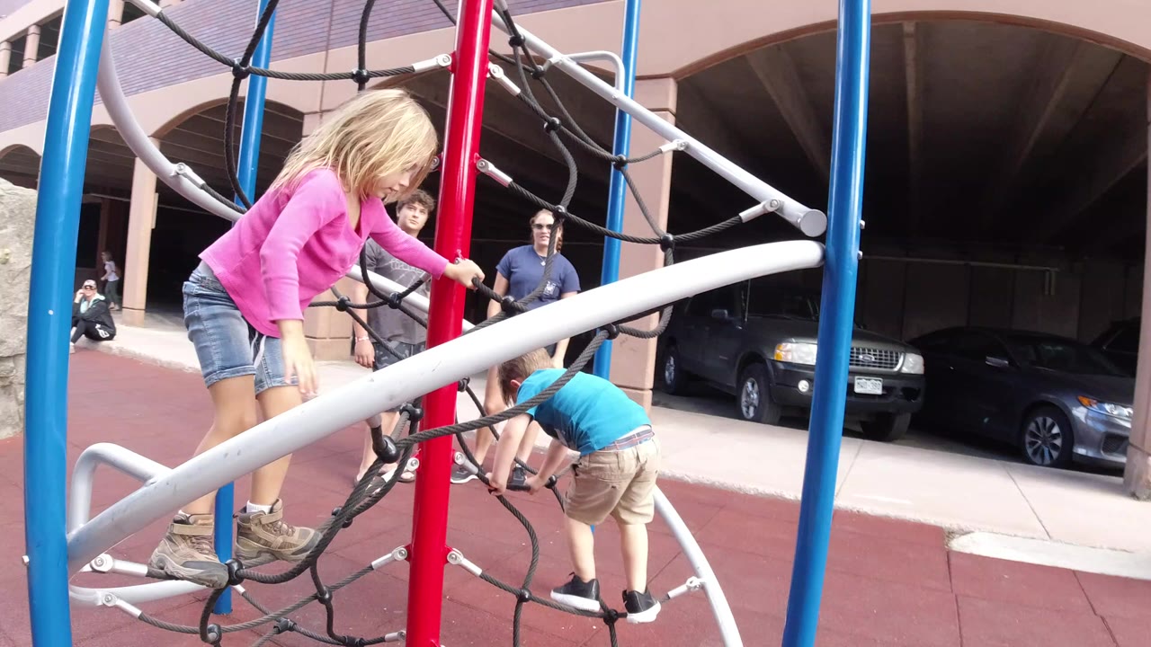 At 0:00 in Winter Park, Colorado, a blonde girl in a pink shirt steps on a black rope net while a younger child in blue bends near the lower ropes and adults stand behind the jungle gym.