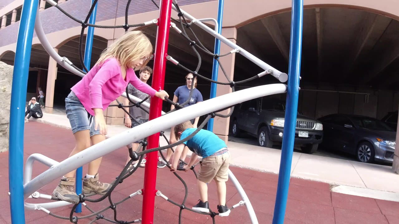 At 0:01 in Winter Park, Colorado, the girl in pink leans toward the red pole on the jungle gym while the child in blue stays low on the ropes and parked cars remain visible under the building archway.