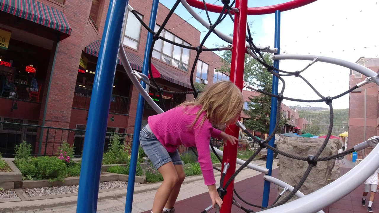 At 0:01 in Winter Park, Colorado, the girl bends forward and reaches toward the black rope net while the red pole, blue posts, silver rail, and a small child in the background remain visible.