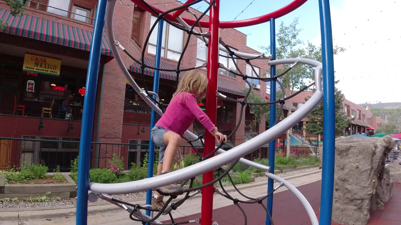 Girl climbing playground ropes beside brick storefronts.