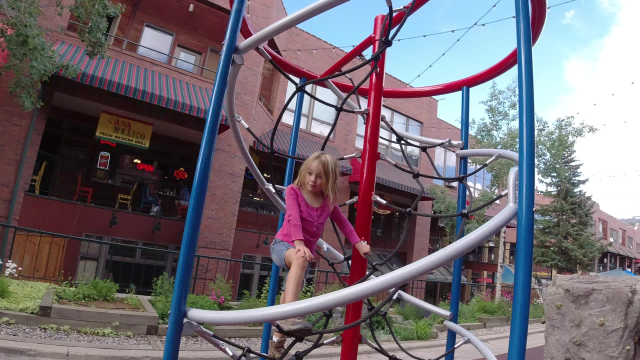 Young girl climbing a playground rope structure near brick storefronts.