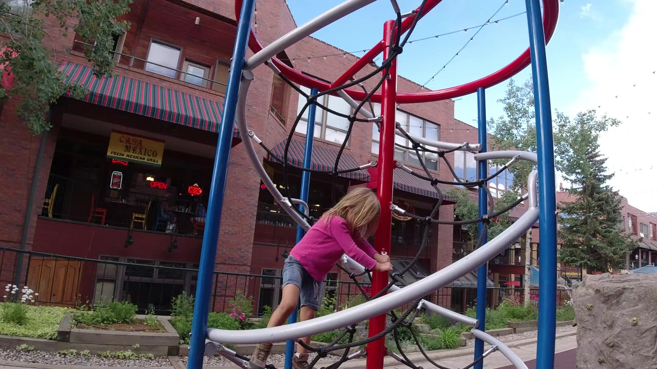 Blonde child climbing a rope play structure near brick buildings.