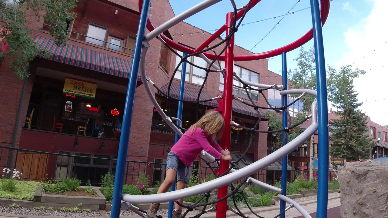 The child continues climbing the rope toy in Winter Park, Colorado, with her body angled forward around the red center pole and silver rail.